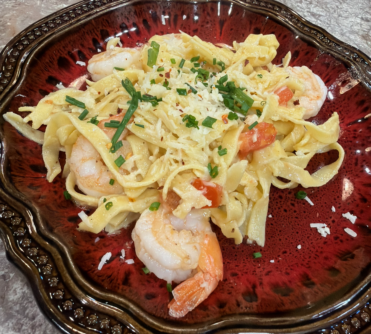Gluten-free homemade fettucine with shrimp in extra-virgin olive oil, plated on a deep red ceramic dish with chives, parmesan, and red pepper flakes